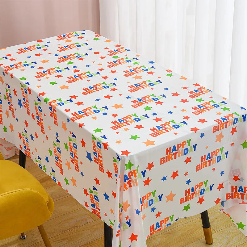 A white disposable plastic tablecloth with a colorful birthday pattern is spread on a table.