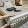 A luxury fitted bed sheet in a beige color is being adjusted on a bed, with a watch and decorative items nearby.