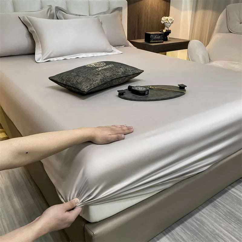 A luxury fitted bed sheet in a beige color is being adjusted on a bed, with a watch and decorative items nearby.