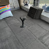A gray plush interlocking puzzle rug mat is displayed on a floor next to a window ledge.