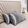 A close-up of a silver satin pillowcase with a quilted headboard and a white lamp on a bedside table.