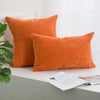 An orange corduroy striped throw pillow cover resting on a white surface next to a green plant.