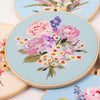 A round embroidery hoop featuring a detailed floral pattern of pink, purple, and white roses and wildflowers on a light blue 