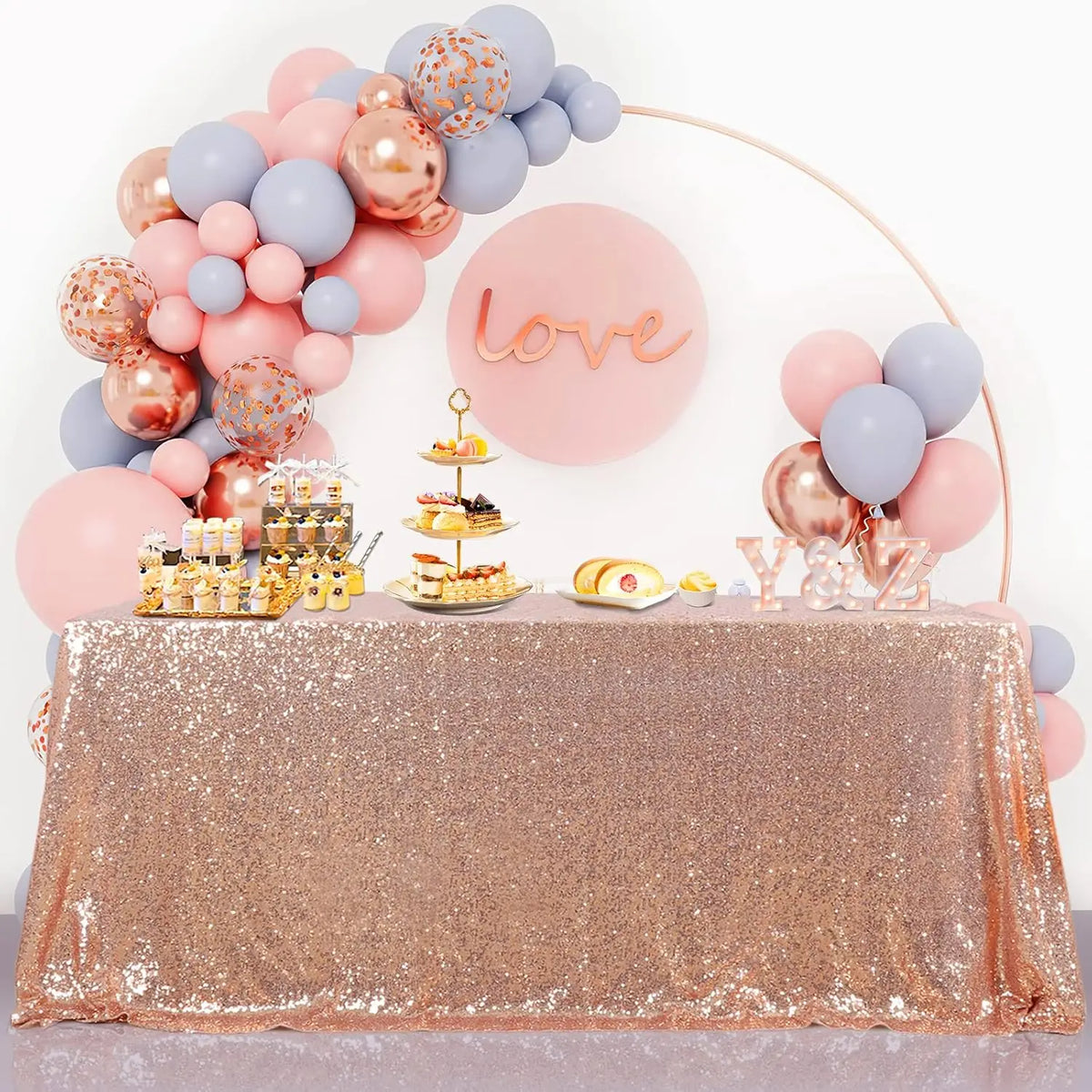 A pink and blue balloon decoration setup with a 'love' sign and a gold tiered cake on a sequin table cover.