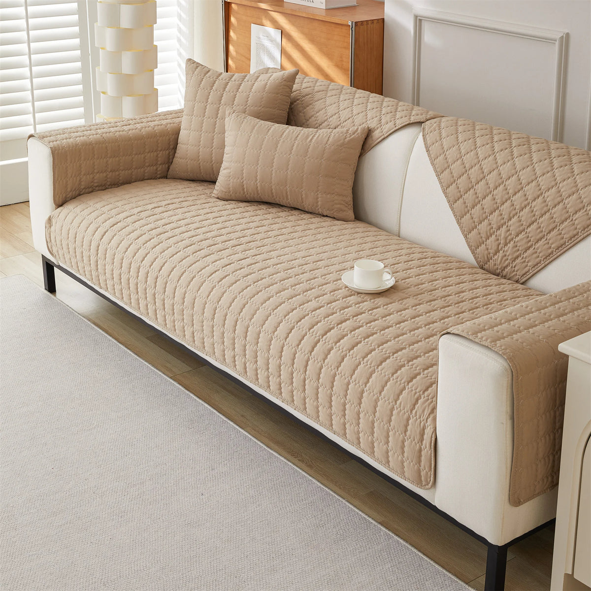 A beige quilted anti-slip sofa cover with plush texture on a white sofa in a bright room.