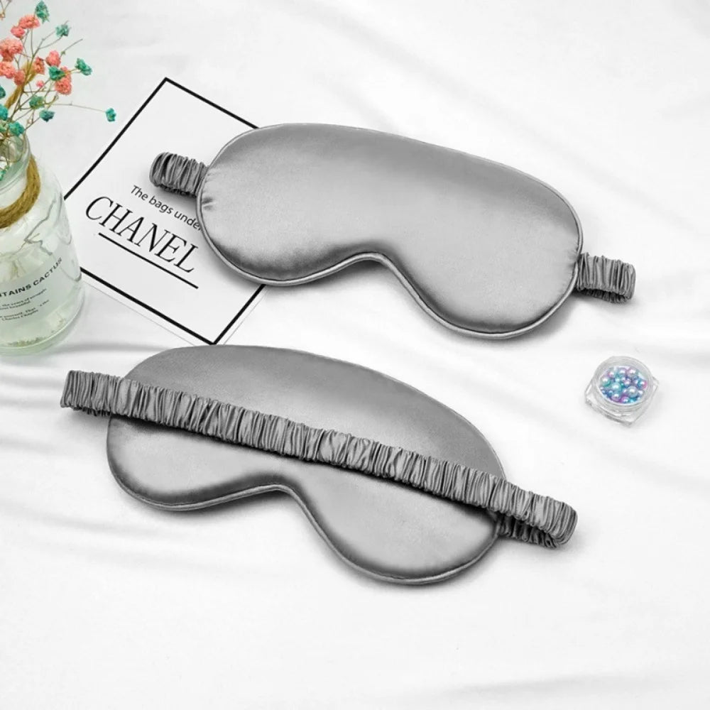 A gray silk sleep mask with a fringe detail is displayed on a white background next to a box labeled CHANEL.