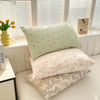 A stack of floral printed pillow covers in pale green and pink tones resting on a white surface near a window.