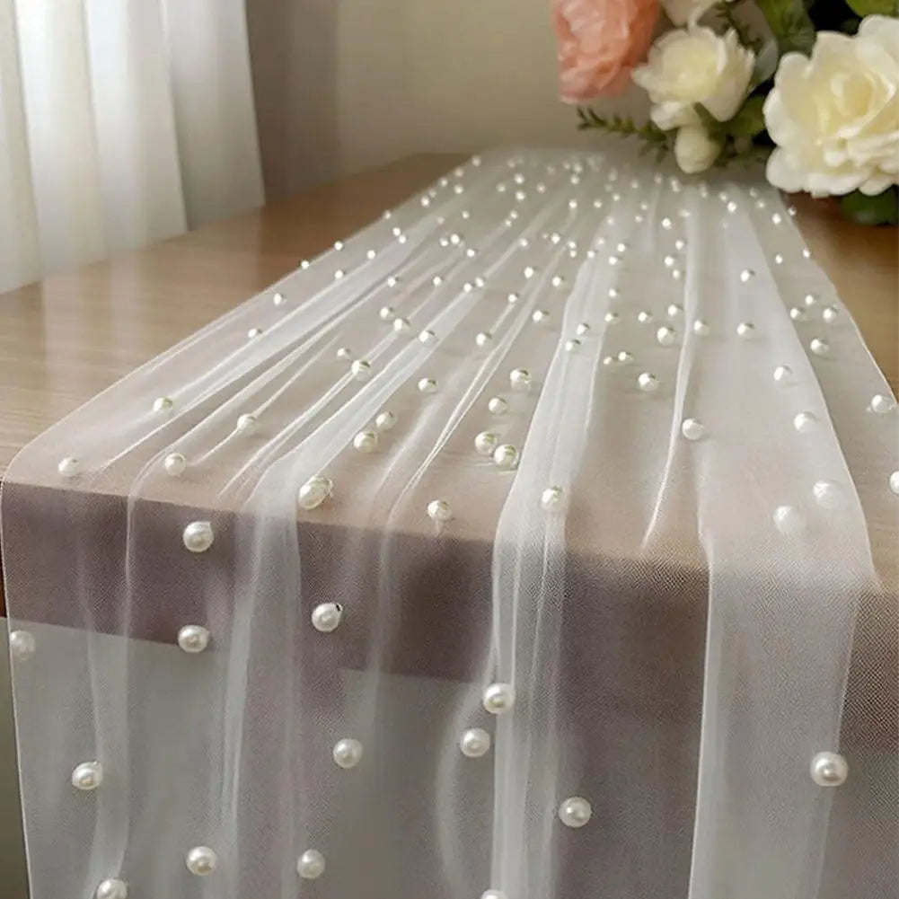 A sheer white tulle table runner adorned with small pearl beads and draped elegantly on a wooden surface.