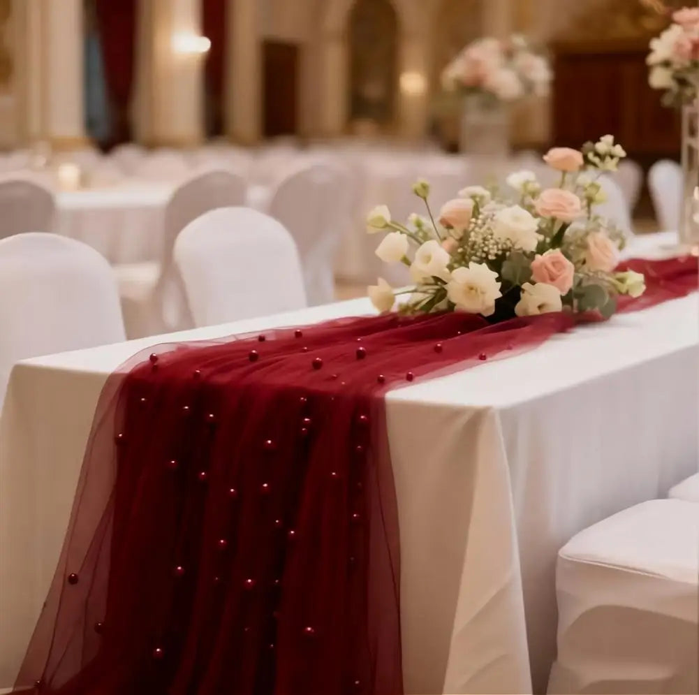 A white table runner with a deep red beaded tulle drape and pink floral arrangement on a white tablecloth.