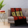 A knitted throw blanket with large orange and yellow letters on a black background draped over a gray sofa.