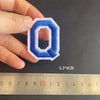 A hand holds a blue embroidered letter Q patch next to a ruler showing its dimensions.