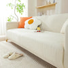 A cream-colored stretchable sofa cover with a vertical line pattern, featuring an orange pillow and a white bear decoration.