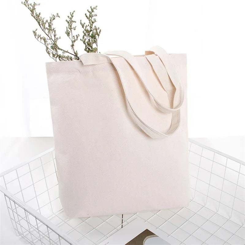 A light beige reusable canvas tote bag with natural twine handles and a sprig of greenery, placed in a white wire basket.