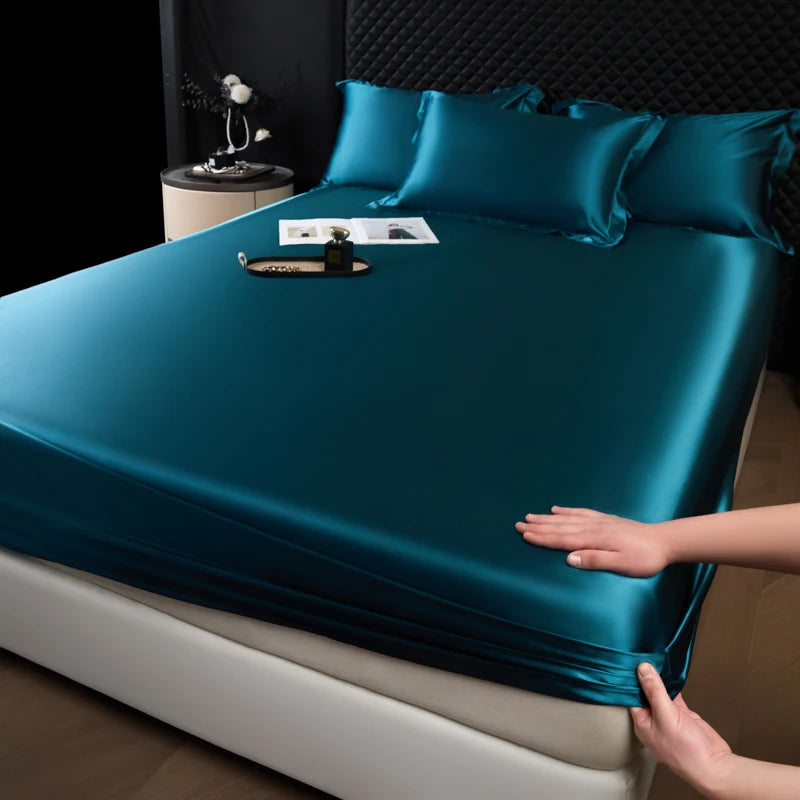 A teal satin fitted sheet is draped over a mattress, with pillows and decorative items visible on the bed.