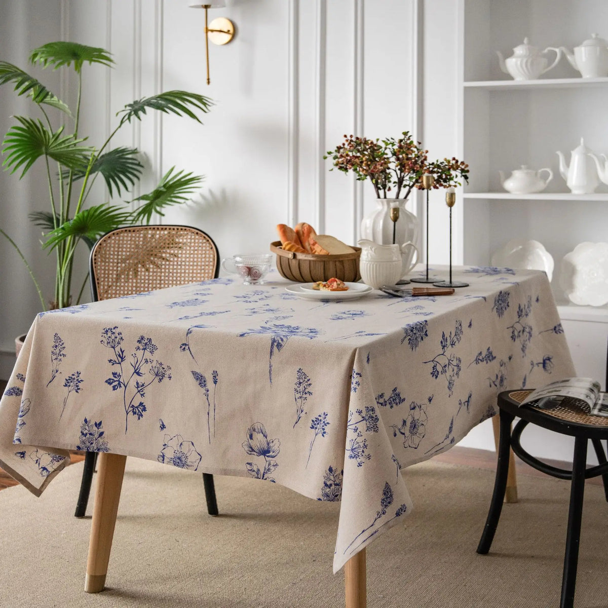 A beige botanical print linen tablecloth is displayed on a wooden table in a bright, modern room.