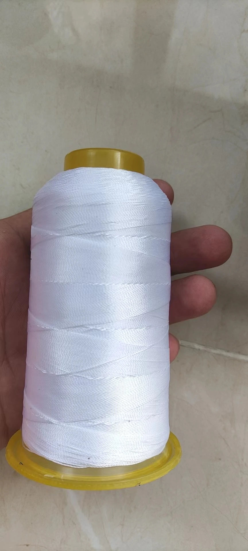 A spool of white high tenacity polyester sewing thread is held in a hand against a light background.