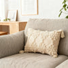 A beige plush pillow cover with a geometric pattern and tassel detail resting on a light gray sofa.