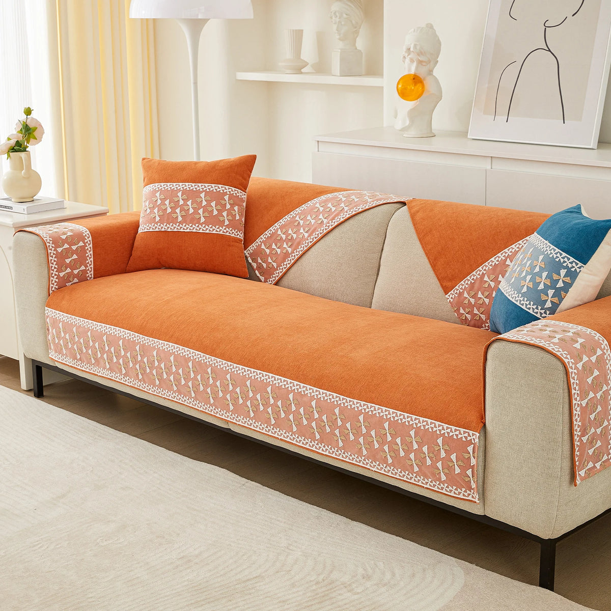 An orange quilted sofa cover with white lace trim and neutral fabric cushions displayed on a light-colored sofa.