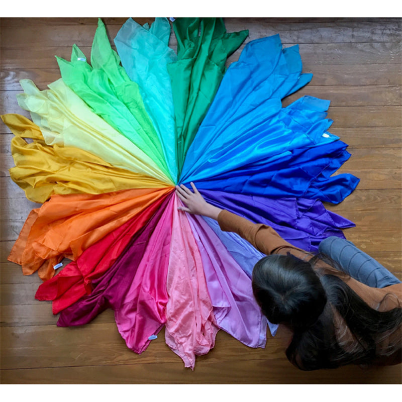 A colorful fan of silk scarves in various shades of green, yellow, blue, purple, and pink laid out on a wooden floor.