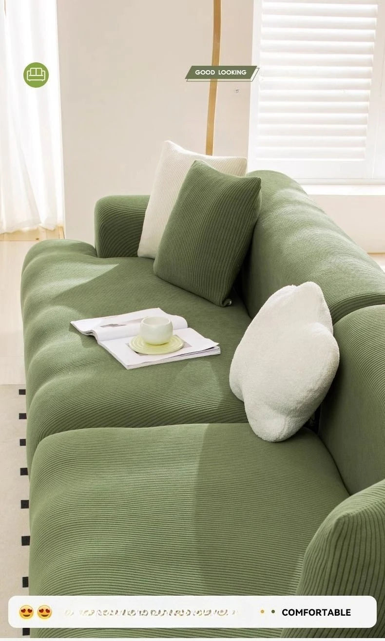 A light green stretchable full-wrap sofa slipcover with white accent pillows and a cup on the seat.