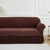 A dark brown jacquard stretch sofa cover with a pleated front draped on a white floor.