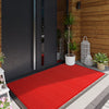 A red non-slip rubber doormat placed on a gray tiled floor in front of a dark wooden door.