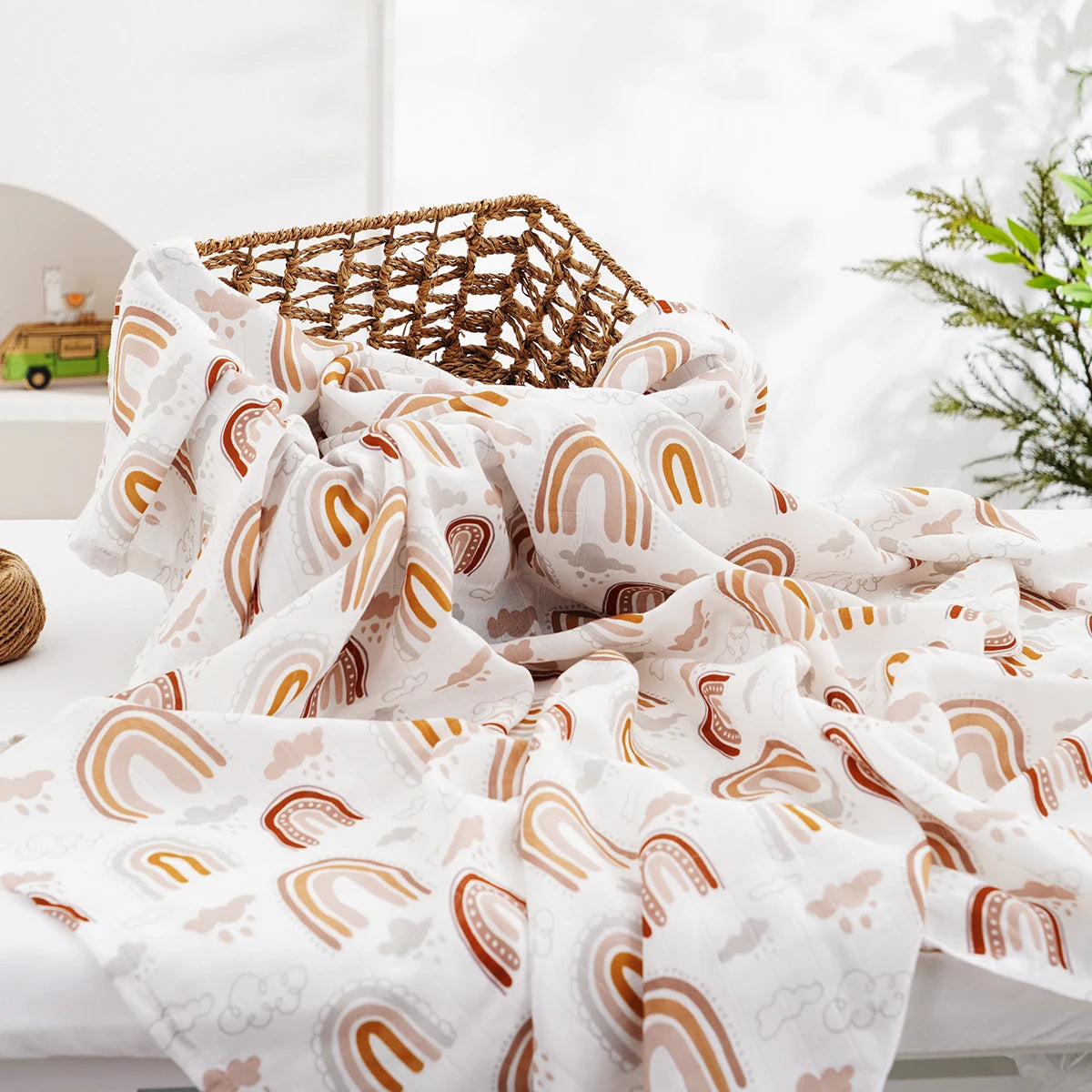 A white muslin swaddle blanket featuring a repeating rainbow pattern in beige and brown tones, draped on a white surface.