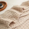 A beige, quilted cotton gauze blanket is draped on a white surface next to a woven basket.