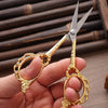 A hand holds a pair of gold-colored crafting scissors with silver blades against a dark wooden background.