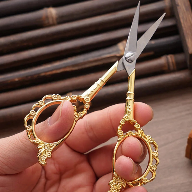 A hand holds a pair of gold-colored crafting scissors with silver blades against a dark wooden background.