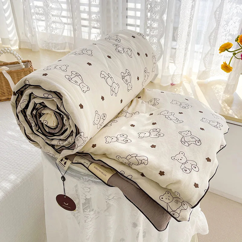 A rolled white quilt with a floral pattern is displayed on a white table covered with a sheer white cloth.