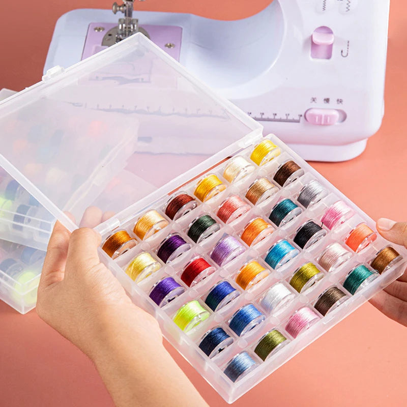 A hand holds an open clear plastic box containing a set of multicolor sewing bobbins and a white sewing machine.
