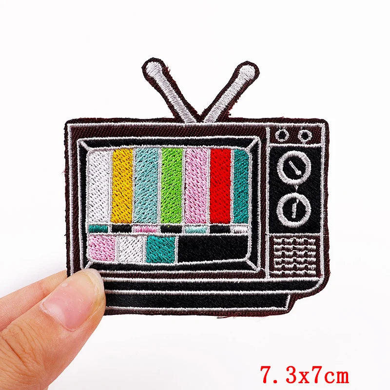 A black embroidered letter patch shaped like an old television set with antenna, featuring colorful vertical stripes.