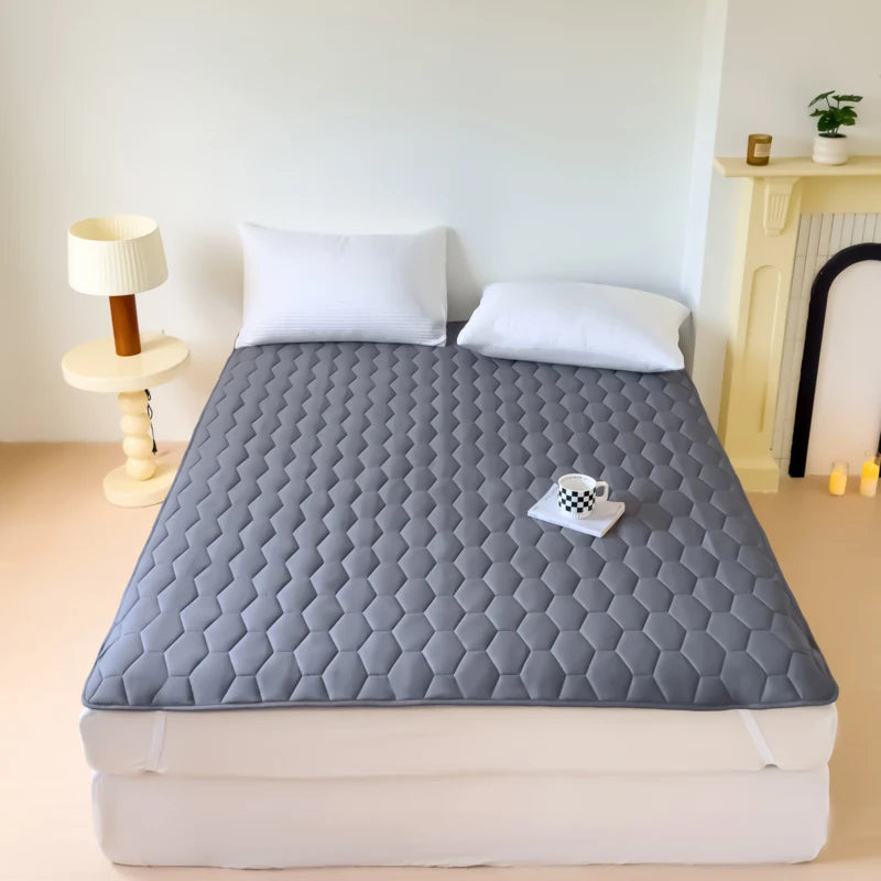 A gray quilted mattress protector cover is displayed on a white bed with a white duvet, next to a lamp and a yellow shelf.