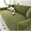 A moss green stretchable full-wrap sofa slipcover with a cream decorative pillow and a cup on the seat.