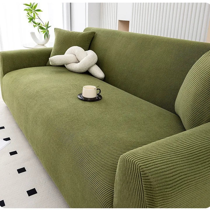 A moss green stretchable full-wrap sofa slipcover with a cream decorative pillow and a cup on the seat.