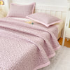 A pink floral quilted bedspread with a pink trim is displayed on a bed with white headboard and pillows.