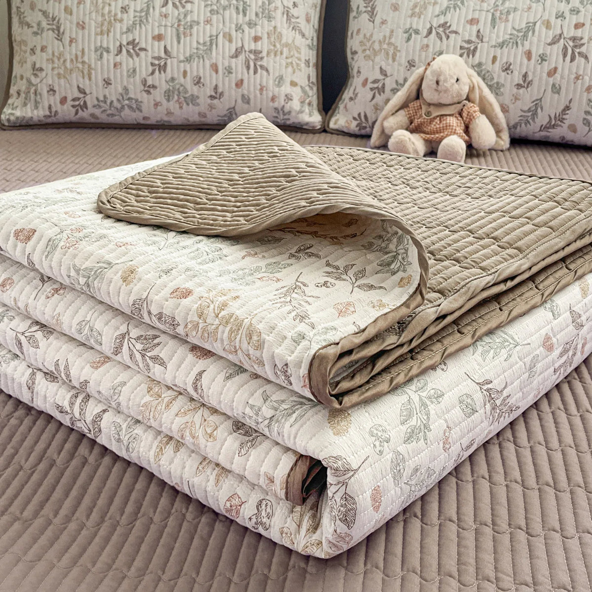 A stack of luxury floral quilted bedspreads in beige and cream tones with a small stuffed rabbit nearby.