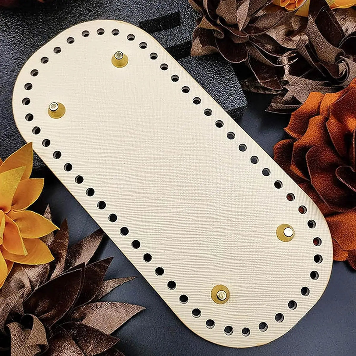 A beige reusable PU leather bag bottom pad with silver grommets rests on a dark surface surrounded by dried orange and brown 