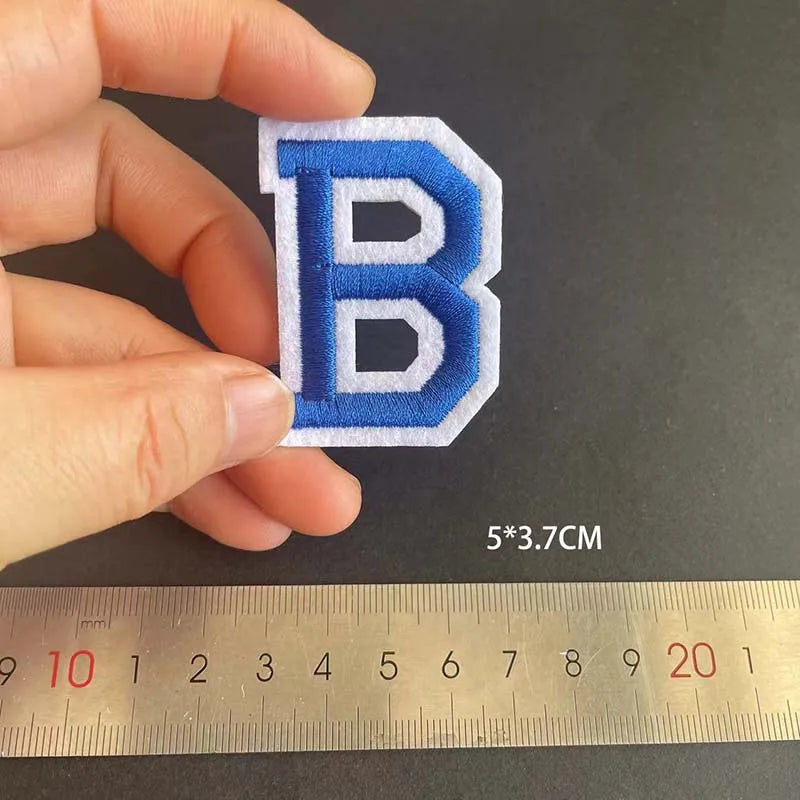 A hand holds a blue embroidered letter B iron-on patch against a ruler.