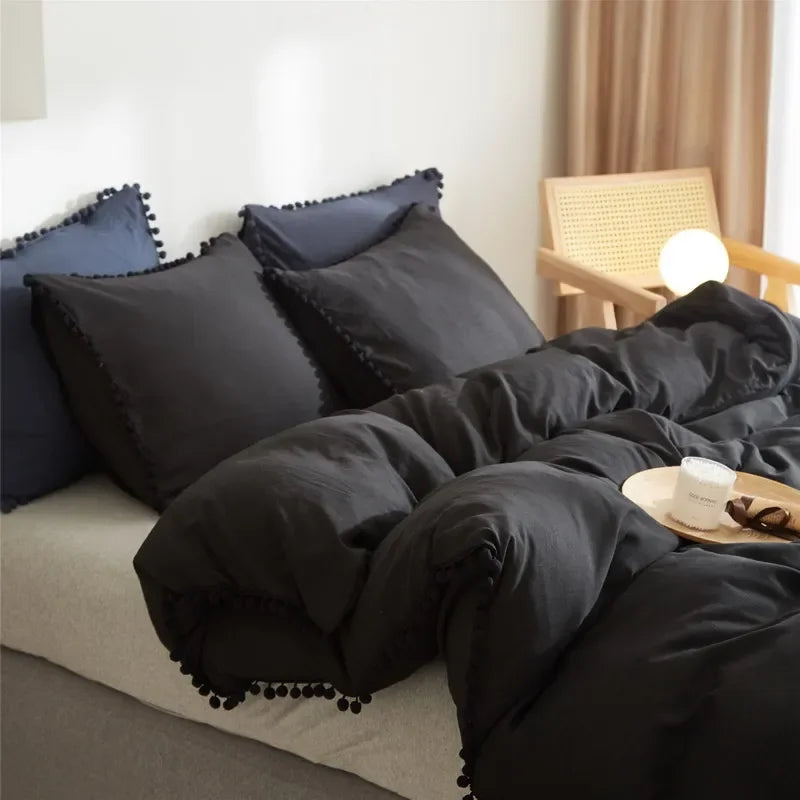 A set of soft washed duvet covers in black and navy blue with pom-pom decoration on a bed.