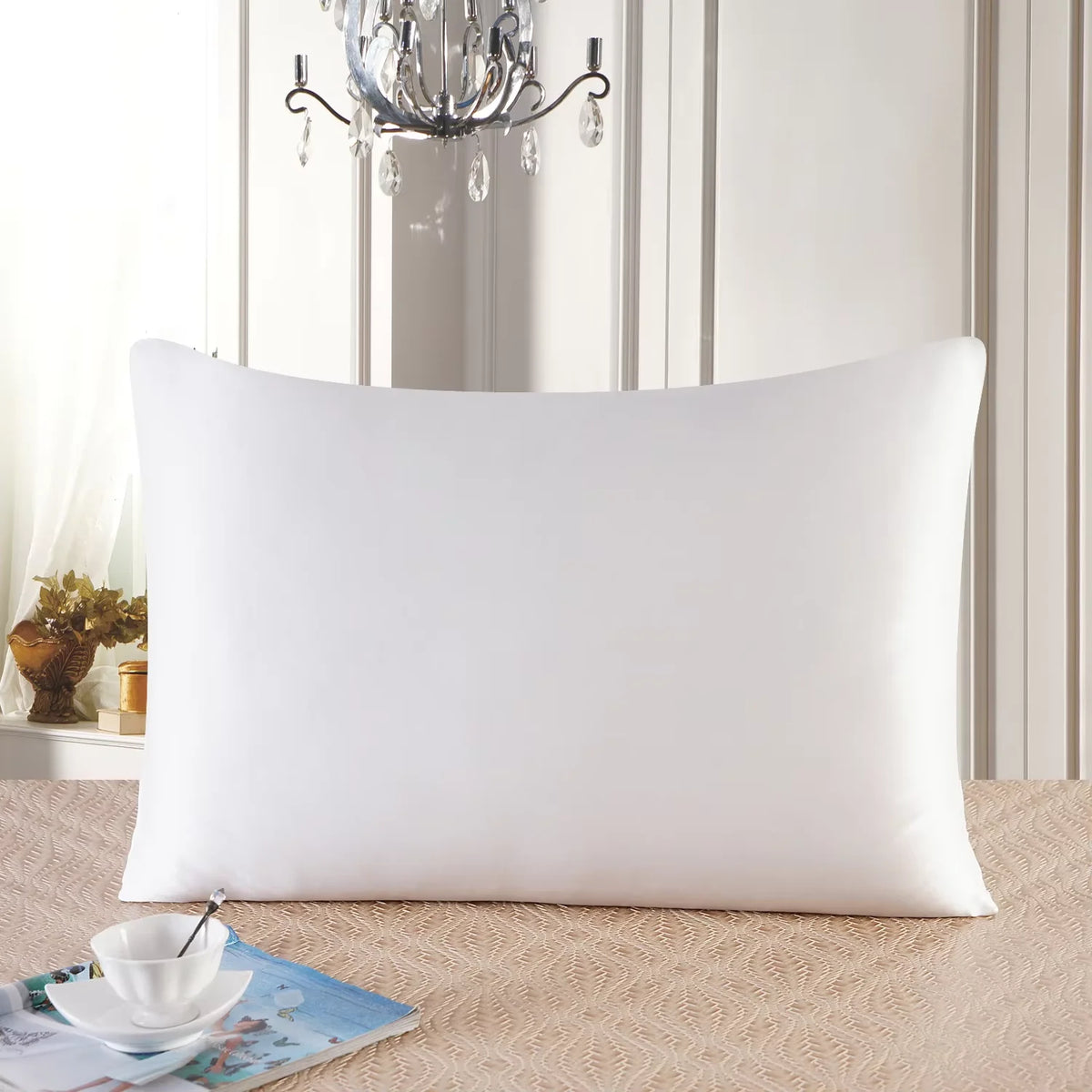 A white pure mulberry silk pillowcase resting on a beige surface in front of a light-colored wall and chandelier.