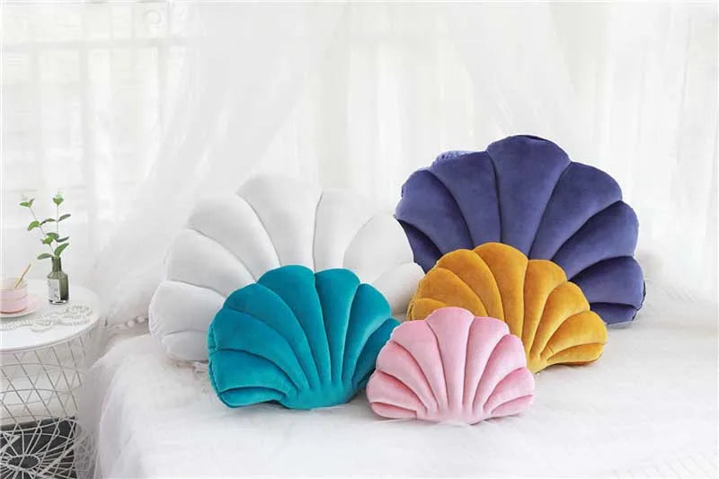 A collection of velvet stuffed sofa cushions shaped like decorative seashells in shades of purple, white, yellow, and teal.