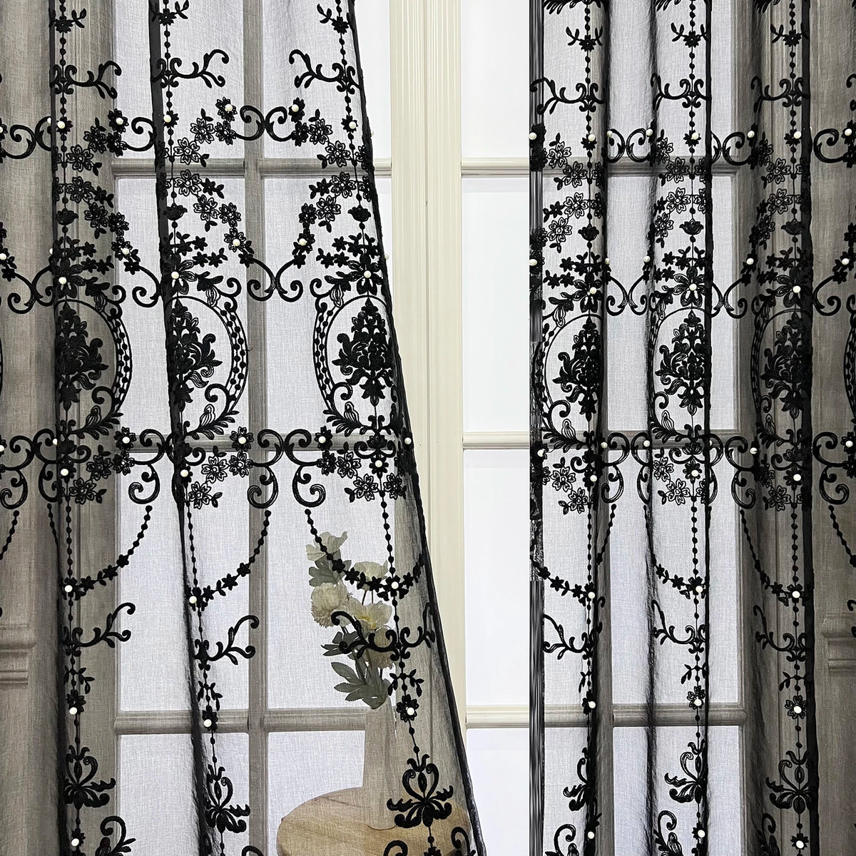 Black sheer curtain featuring intricate lace embroidery and pearl detailing hanging in a window.