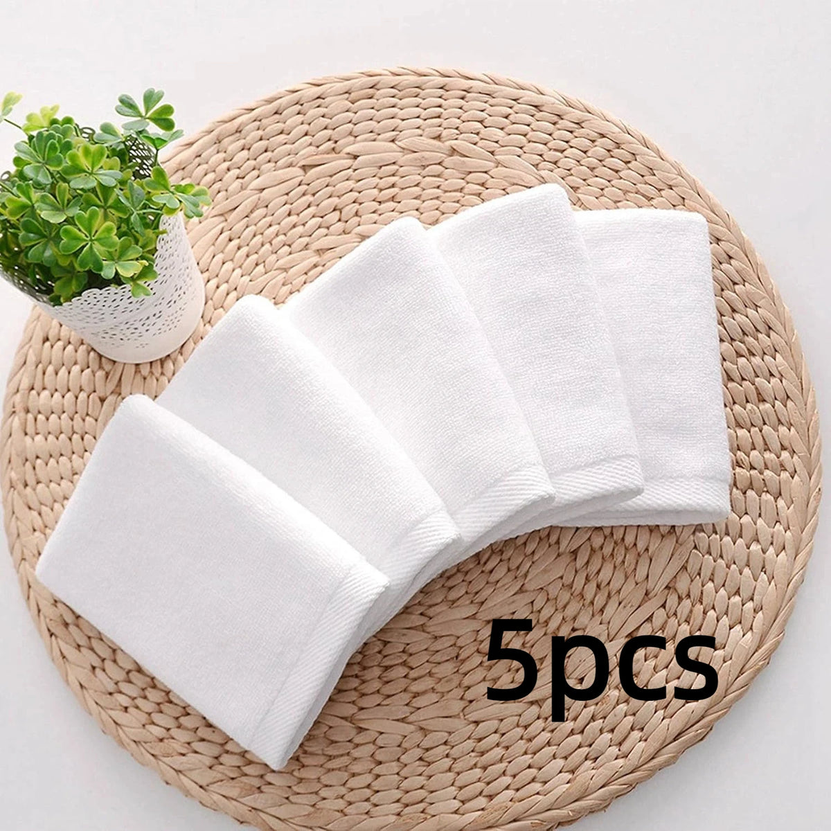 A stack of five white cotton face towels resting on a woven mat next to a small potted plant.
