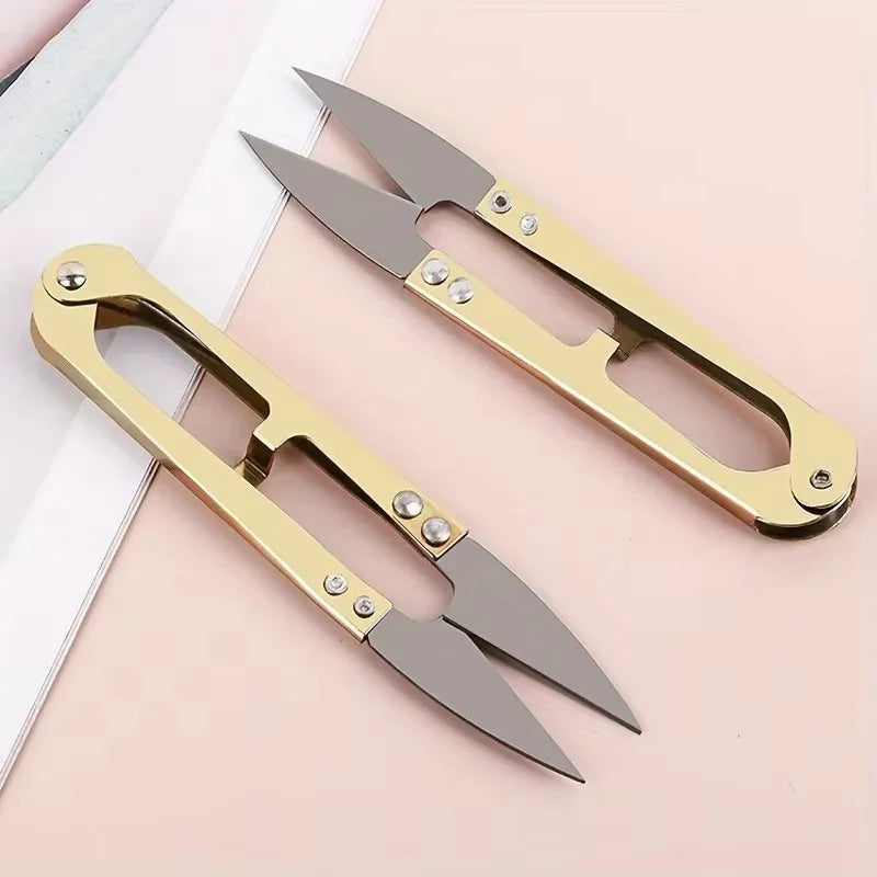 A pair of gold-colored U-shape sewing scissors with sharp blades resting on a light pink surface.