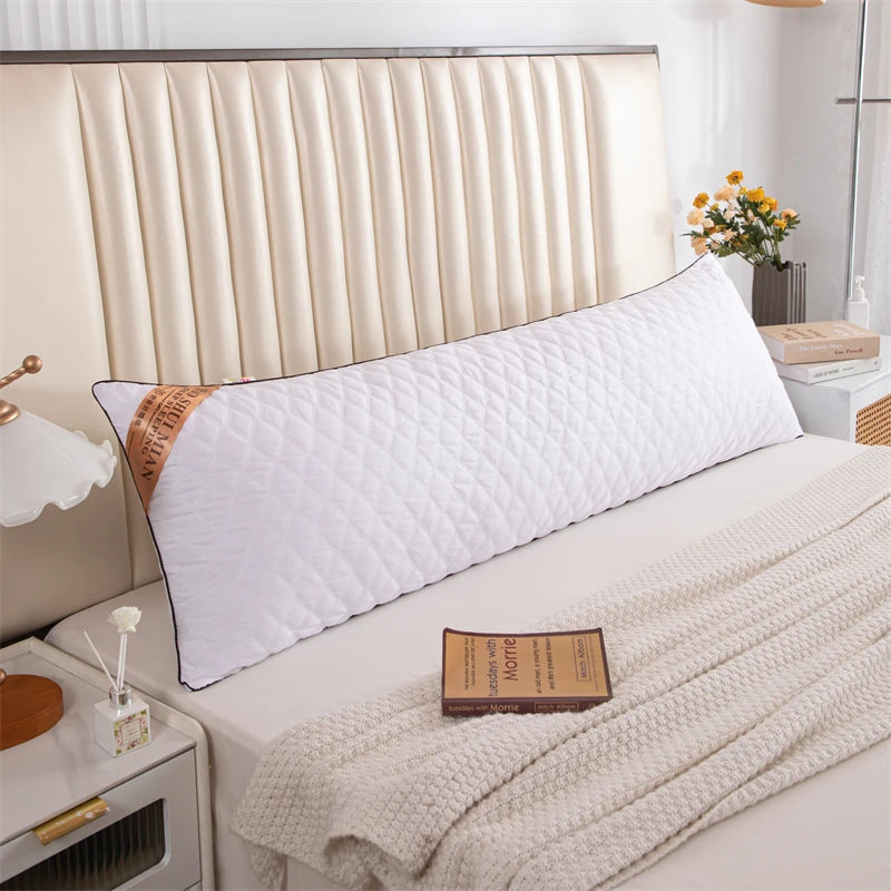 A white quilted rectangle sleeping pillow rests on a bed with a light beige headboard and a cream blanket.