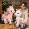A woman is holding a white unicorn plush toy with rainbow accents, sitting next to a laptop.