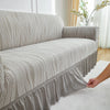 A beige jacquard stretch sofa cover draped over a light gray sofa, showing pleats and gathered fabric.