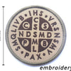 A circular embroidered patch featuring various letters and symbols in dark brown thread on a tan background.